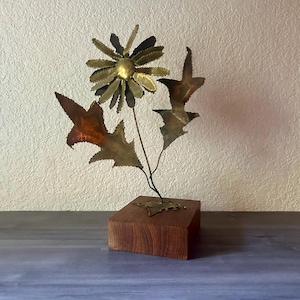 May include: A handcrafted metal flower sculpture with a brass center and petals, copper leaves, and a wooden base. The flower has a detailed design and is a unique decorative piece.
