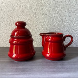 May include: A red ceramic sugar bowl with a lid and a matching creamer. The sugar bowl has a rounded shape with a lid and a small handle. The creamer has a handle and a spout. Both pieces have a glossy finish.