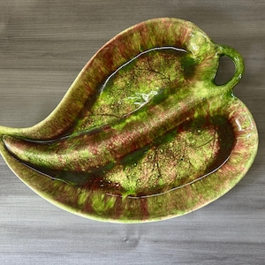 May include: A leaf-shaped ceramic serving dish with a handle. The dish features a glossy finish with a blend of green, brown, and yellow hues. The design includes a textured leaf pattern, adding a naturalistic touch to the serving piece.