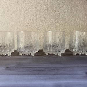 May include: A set of four clear glass tumblers featuring a textured, bark-like exterior pattern and small, rounded feet at the base. Each glass has a cylindrical shape with a consistent vertical design and a smooth, transparent finish.