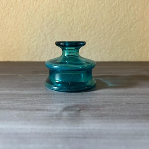 May include: A teal-colored glass bottle stopper with a rounded base and a flared top. The stopper is made of transparent glass and sits on a gray wooden surface. The background is a light beige wall.