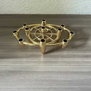 May include: A gold-colored metal candle holder with space for ten candles. The candle holders are black. The design is circular with a central wheel-like structure and three supporting legs.