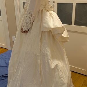 May include: A long-sleeved ivory gown featuring intricate white lace and beadwork on the bodice and sleeves. The dress includes a fitted waist with a decorative fabric flower accent and a full, floor-length skirt with a layered, ruffled bustle.
