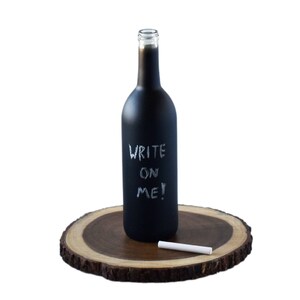 Peut inclure: Une bouteille de vin en verre noir avec une finition en ardoise. La bouteille porte l'inscription "WRITE ON ME!" écrite à la craie blanche. Un morceau de craie est posé sur une tranche de bois à côté de la bouteille.