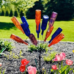 May include: A cluster of colorful glass bottle garden art in shades of orange, blue, and purple. The repurposed bottles are mounted on metal stakes, creating a unique outdoor decoration. The garden art is surrounded by flowers and greenery.