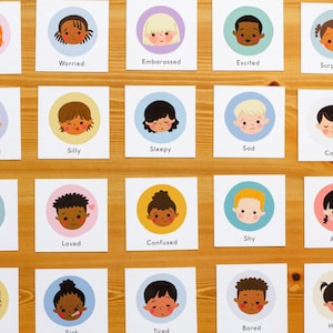 May include: A collection of emotion flashcards featuring cartoon faces. Each card displays a different emotion, such as "Scared," "Worried," and "Happy." The cards have a white background with a colored circle around each face. The cards are arranged on a wooden surface.