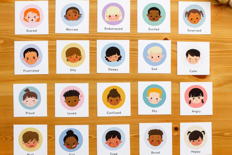 May include: A collection of emotion flashcards featuring cartoon faces. Each card displays a different emotion, such as "Scared," "Worried," and "Happy." The cards have a white background with a colored circle around each face. The cards are arranged on a wooden surface.