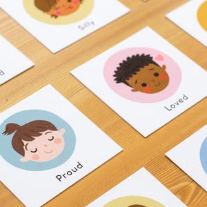 May include: Cards with illustrations of diverse children's faces expressing emotions like frustration, silliness, and love. Each card features a different emotion label, such as "Proud" or "Loved", set against a colored circle background. The cards are arranged on a wooden surface.