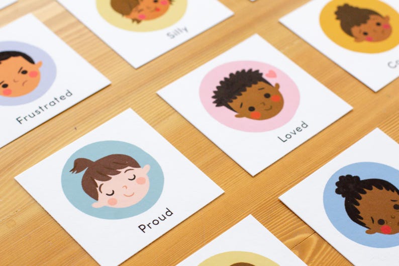May include: Cards with illustrations of diverse children's faces expressing emotions like frustration, silliness, and love. Each card features a different emotion label, such as "Proud" or "Loved", set against a colored circle background. The cards are arranged on a wooden surface.