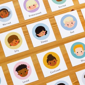 May include: Cards with illustrations of children's faces expressing different emotions, such as worried, embarrassed, and excited. Each card features a circular illustration on a white background, with the emotion's name below. The cards are arranged on a wooden surface.
