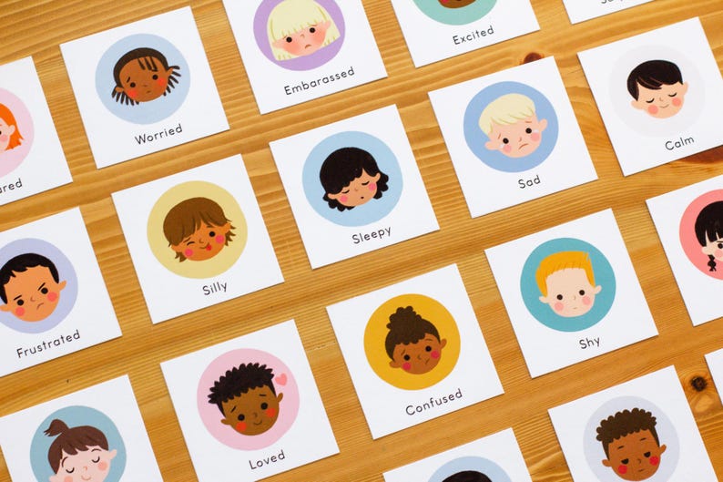 May include: Cards with illustrations of children's faces expressing different emotions, such as worried, embarrassed, and excited. Each card features a circular illustration on a white background, with the emotion's name below. The cards are arranged on a wooden surface.