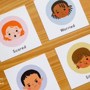 May include: Cards with illustrations of children's faces expressing emotions. The cards include the words "Scared", "Worried", and "Silly". The phrase "illustrations made by hand" is also visible. The cards are on a wooden surface.