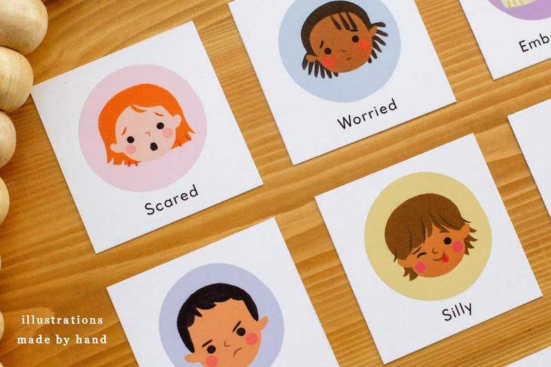May include: Cards with illustrations of children's faces expressing emotions. The cards include the words "Scared", "Worried", and "Silly". The phrase "illustrations made by hand" is also visible. The cards are on a wooden surface.