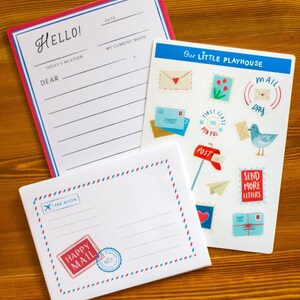May include: A collection of stationery items, including a notepad with "Hello!" and lined sections, a sheet of stickers with mail-themed illustrations, and a postcard with "Happy Mail" text. The items are arranged on a wooden surface.