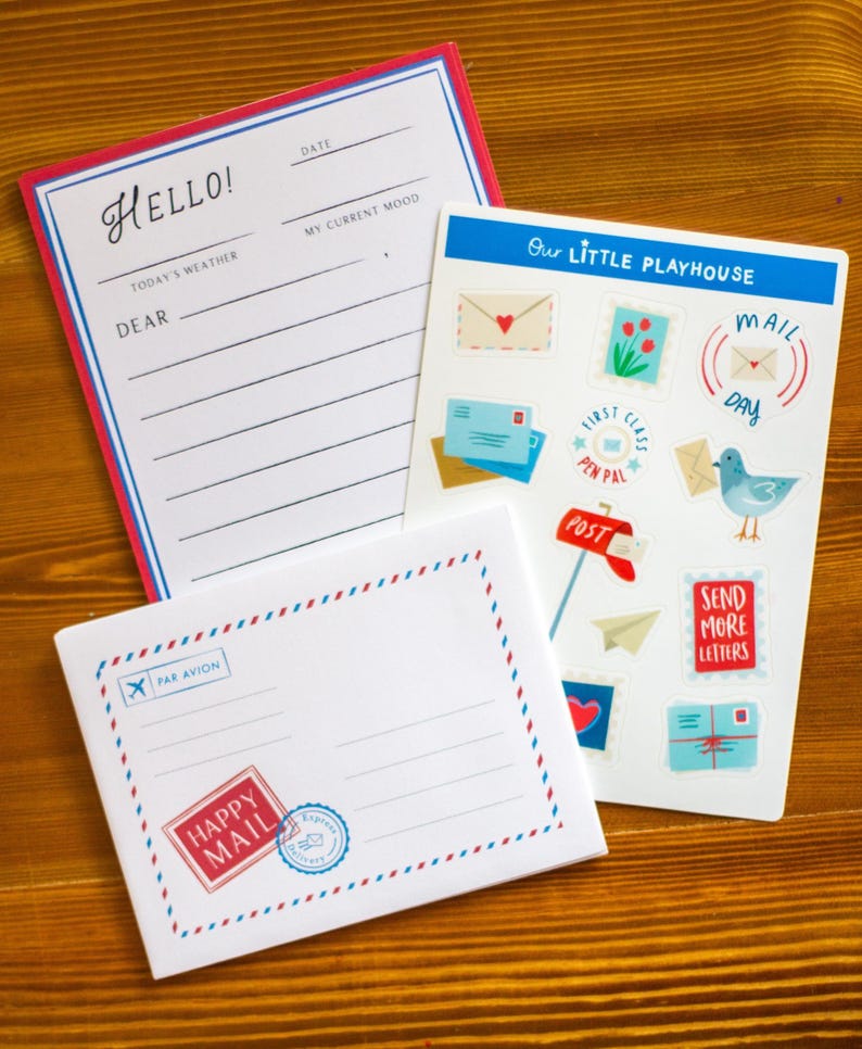 May include: A collection of stationery items, including a notepad with "Hello!" and lined sections, a sheet of stickers with mail-themed illustrations, and a postcard with "Happy Mail" text. The items are arranged on a wooden surface.