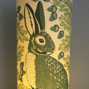 May include: A green and white lampshade with a rabbit and acorns design. The lampshade is lit from the inside, casting a warm glow.