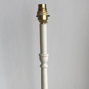 May include: A cream-coloured lamp base with a brass bulb socket. The base has a round, tiered bottom and a slender, turned column. The lamp base is approximately 30 cm tall.