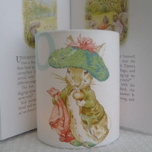 May include: A white candle holder featuring a printed image of a rabbit wearing a green and blue hat and holding a carrot. The text "UNFORTUN time a floc from bush to caterpillars a several sorts different songs The first digging-upmy up my nuts?" is printed on the left side of the candle holder.