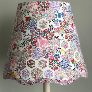 May include: A lampshade with a floral pattern and a scalloped edge. The design is a patchwork of hexagonal shapes filled with flowers in pink, blue, red, and yellow. The base is a light cream colour.