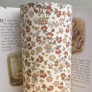 May include: Cylindrical lampshade with a floral pattern in shades of brown, cream, and yellow. The design includes small figures and is set against a cream background. The lampshade is placed in front of an open book.