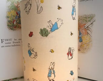 May include: A white cylindrical lampshade with a repeating pattern of Peter Rabbit characters, including Peter Rabbit, a squirrel, bees, and butterflies. The lampshade is lit from within, casting a warm glow.