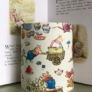 Op de afbeelding: Cilindrische lampenkap in cr&egrave;me met een grillige illustratie van muizen, eekhoorns en voedsel zoals cupcakes en taarten. Het ontwerp heeft een sprookjesachtige uitstraling. De lampenkap staat voor een open boek.