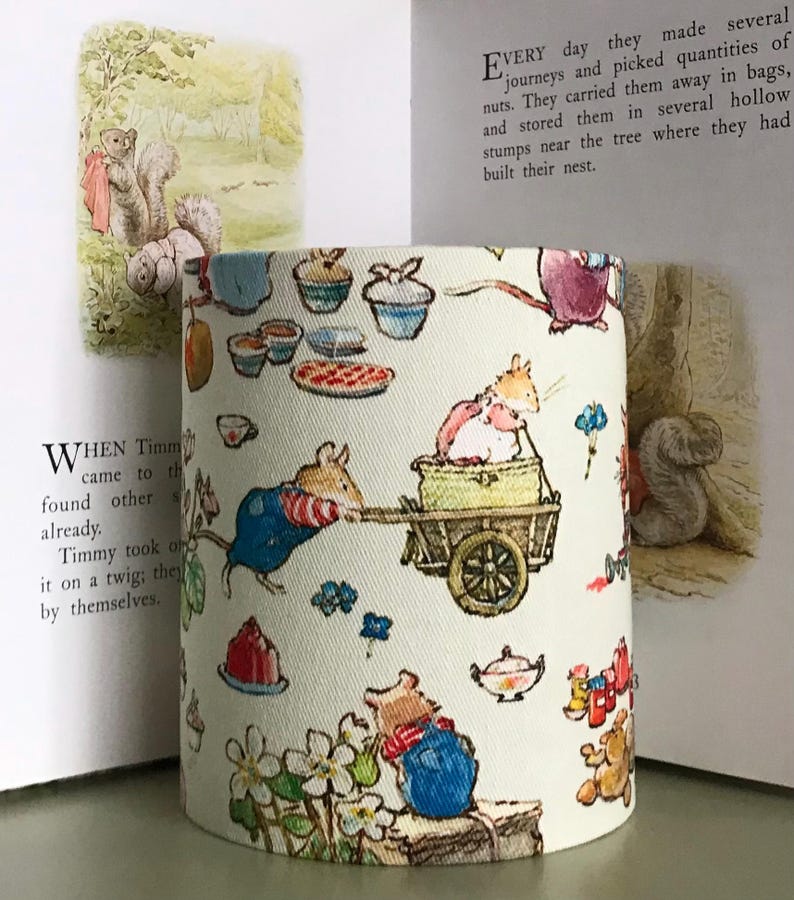 Op de afbeelding: Cilindrische lampenkap in cr&egrave;me met een grillige illustratie van muizen, eekhoorns en voedsel zoals cupcakes en taarten. Het ontwerp heeft een sprookjesachtige uitstraling. De lampenkap staat voor een open boek.