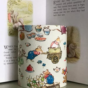 Op de afbeelding: Cilindrische lampenkap met een grillige illustratie van muizen, theekopjes en gebak. Het ontwerp bevat een muis die een kar trekt en andere muizen in overalls. De achtergrond is lichtcr&egrave;me van kleur, met tekst uit een kinderboek zichtbaar.