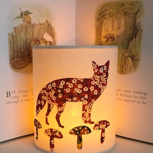May include: A white lampshade featuring a red fox silhouette printed on it. The fox is surrounded by small white flowers and is standing on top of three mushrooms. The lampshade is lit from within, casting a warm glow.