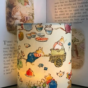 Op de afbeelding: Cilindrische lampenkap met een grillige illustratie van muizen, eten en bloemen. Het ontwerp bevat muizen die een kar trekken en van lekkernijen genieten. De achtergrond toont pagina's uit een kinderboek, wat een gezellige, sprookjesachtige esthetiek cre&euml;ert.