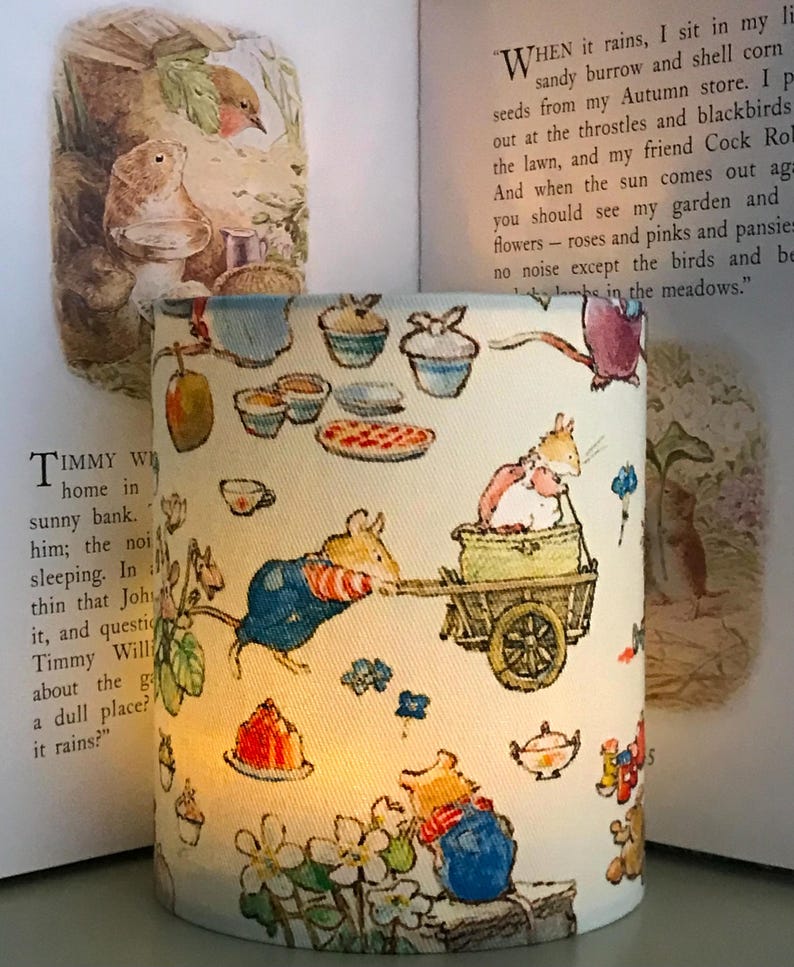 Op de afbeelding: Cilindrische lampenkap met een grillige illustratie van muizen, eten en bloemen. Het ontwerp bevat muizen die een kar trekken en van lekkernijen genieten. De achtergrond toont pagina's uit een kinderboek, wat een gezellige, sprookjesachtige esthetiek cre&euml;ert.