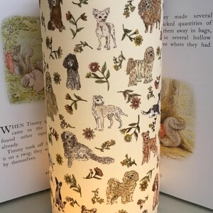 May include: Cylindrical lampshade with a cream background, featuring illustrations of various dog breeds and floral accents. The design includes a variety of dog breeds, flowers, and foliage. The lampshade is placed between two open books.