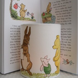 May include: A white ceramic candle holder featuring a Winnie the Pooh theme. The holder depicts illustrations of Winnie the Pooh, Piglet, Rabbit, and Kanga. The illustrations are in a cartoon style and are coloured in shades of brown, yellow, pink, and green. The candle holder is cylindrical in shape and has a green base.