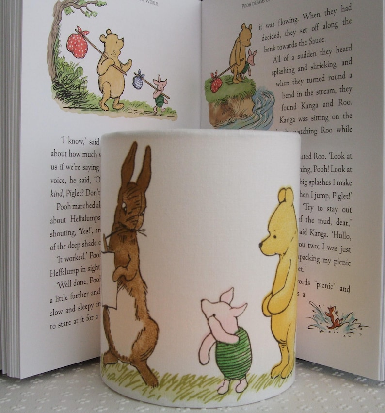 May include: A white ceramic candle holder featuring a Winnie the Pooh theme. The holder depicts illustrations of Winnie the Pooh, Piglet, Rabbit, and Kanga. The illustrations are in a cartoon style and are coloured in shades of brown, yellow, pink, and green. The candle holder is cylindrical in shape and has a green base.