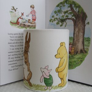 May include: A white ceramic mug featuring a Winnie the Pooh design. The mug depicts Winnie the Pooh, Piglet and Rabbit. The mug is decorated in a green, brown and yellow colour scheme. The mug is resting against a book with text about Winnie the Pooh.