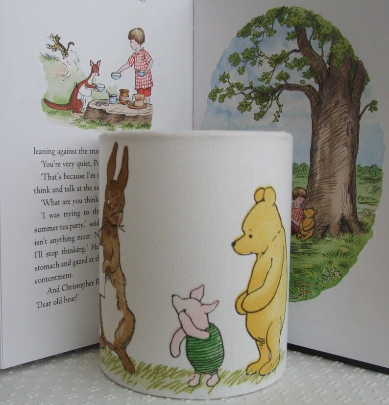 May include: A white ceramic mug featuring a Winnie the Pooh design. The mug depicts Winnie the Pooh, Piglet and Rabbit. The mug is decorated in a green, brown and yellow colour scheme. The mug is resting against a book with text about Winnie the Pooh.