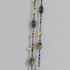 May include: A long silver-toned chain necklace featuring a series of decorative metal links interspersed with small, colorful beads in shades of blue, yellow, pink, and clear, along with ornate, diamond-shaped metal charms with inlaid accents.