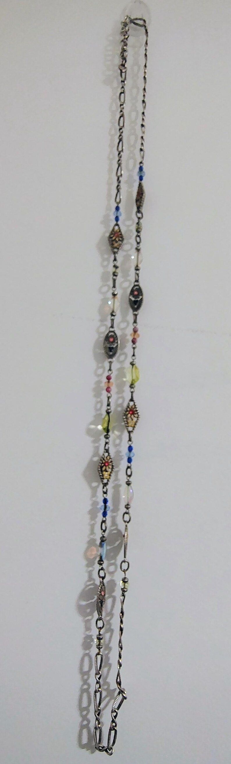 May include: A long silver-toned chain necklace featuring a series of decorative metal links interspersed with small, colorful beads in shades of blue, yellow, pink, and clear, along with ornate, diamond-shaped metal charms with inlaid accents.