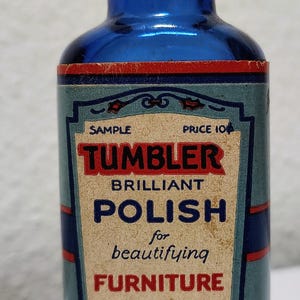 May include: A vintage blue glass bottle of Tumbler Brilliant Polish for furniture. The label reads "Cleans as it Polishes" and "Shines in a jiffy." The bottle has a black cap and a paper label with red and blue accents.