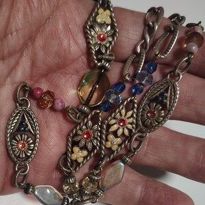 May include: A silver-toned metal chain necklace featuring oval and diamond-shaped filigree links decorated with small cream-colored flowers, red and pink rhinestones, and interspersed with translucent blue, yellow, and iridescent glass beads.
