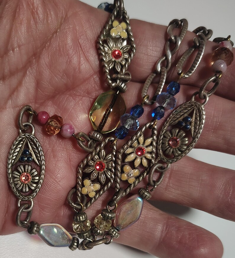 May include: A silver-toned metal chain necklace featuring oval and diamond-shaped filigree links decorated with small cream-colored flowers, red and pink rhinestones, and interspersed with translucent blue, yellow, and iridescent glass beads.