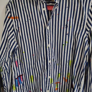 May include: A button-down shirt with vertical navy blue and white stripes. The shirt has colorful paint splatters in orange, pink, yellow, and green. The shirt has a collar and buttoned cuffs. The Polo Ralph Lauren logo is visible on the shirt.
