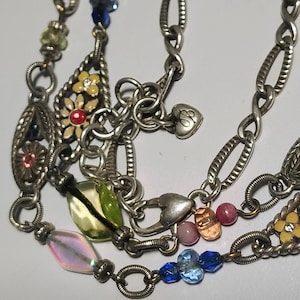 May include: A silver-toned charm necklace featuring multiple oval chain links, small heart-shaped charms, and various decorative elements including yellow flower charms with red centers, iridescent beads, and blue glass beads.
