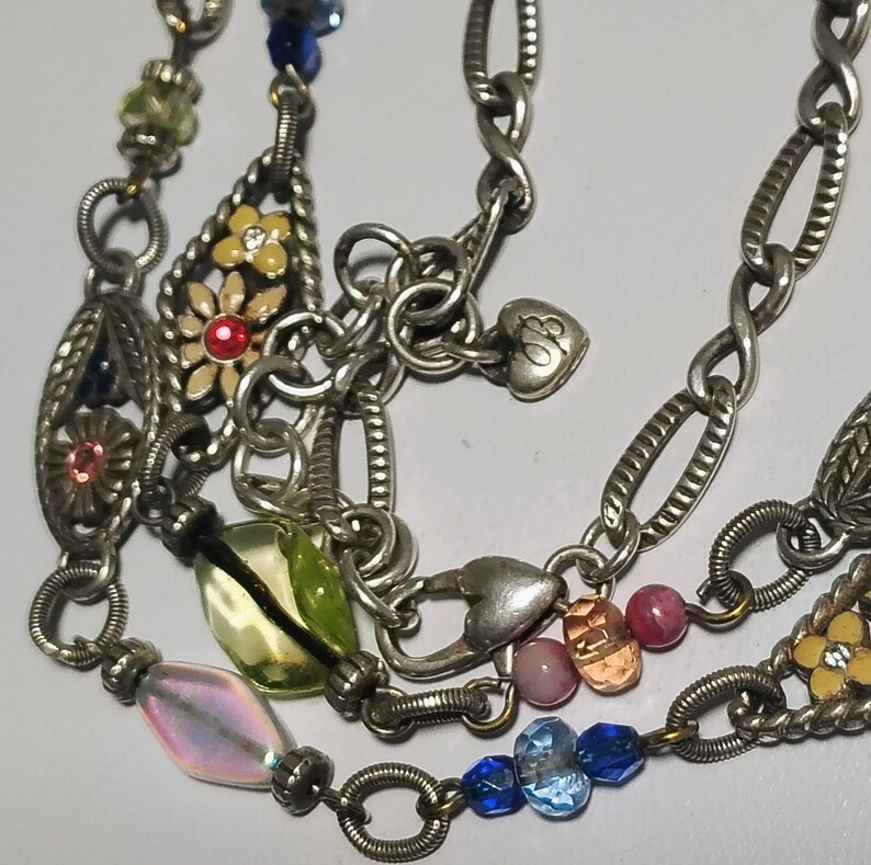 May include: A silver-toned charm necklace featuring multiple oval chain links, small heart-shaped charms, and various decorative elements including yellow flower charms with red centers, iridescent beads, and blue glass beads.