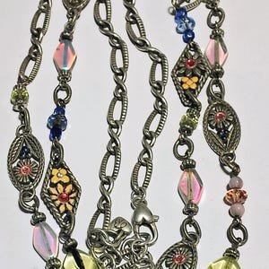 May include: A long silver-tone metal chain necklace featuring oval and diamond-shaped filigree links with floral motifs. The necklace is accented with multi-colored glass beads in shades of blue, pink, yellow, and green, and includes a lobster clasp.