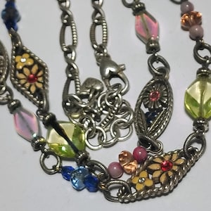 May include: A silver-toned metal chain necklace featuring a series of oval-shaped links with floral motifs in yellow and red enamel, interspersed with translucent glass beads in shades of blue, pink, lime green, and amber, and a lobster clasp.