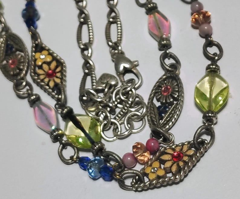 May include: A silver-toned metal chain necklace featuring a series of oval-shaped links with floral motifs in yellow and red enamel, interspersed with translucent glass beads in shades of blue, pink, lime green, and amber, and a lobster clasp.