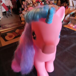 May include: A pink toy pony with blue and purple hair. The pony has blue eyes and a black eye design. The toy is standing on a dark surface. The pony is a children's toy.