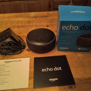 May include: An Amazon Echo Dot smart speaker, power adapter, and packaging are displayed on a wooden surface. The box is blue and white, with the words "echo dot" and "Add Alexa to any room." Two instruction cards are also present.