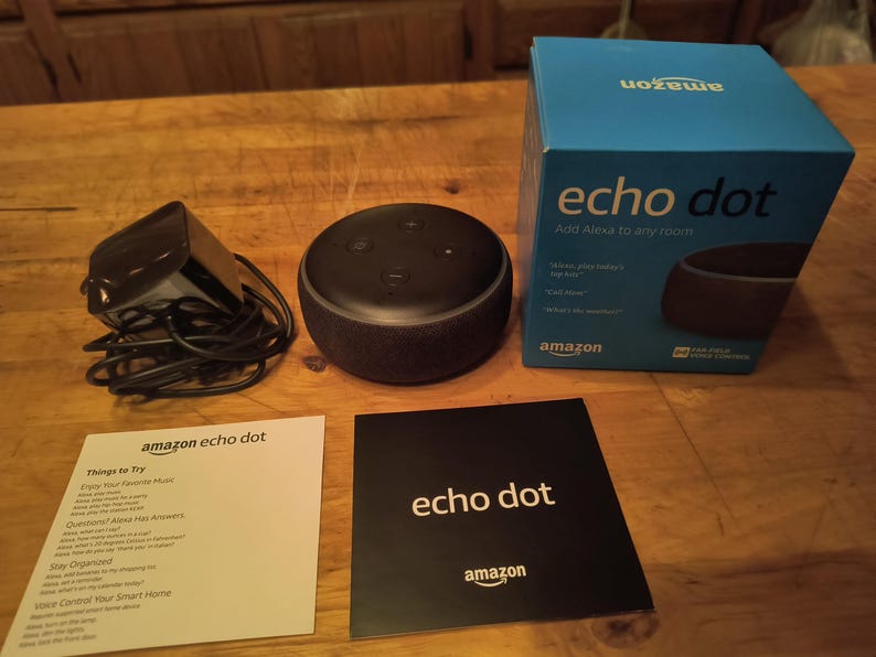 K&ouml;nnte beinhalten: Ein schwarzer Amazon Echo Dot Smart Speaker, Netzteil und Verpackung liegen auf einer Holzoberfl&auml;che. Die blaue Box tr&auml;gt die Aufschrift "echo dot" und "Add Alexa to any room". Zwei Anweisungskarten sind ebenfalls vorhanden.